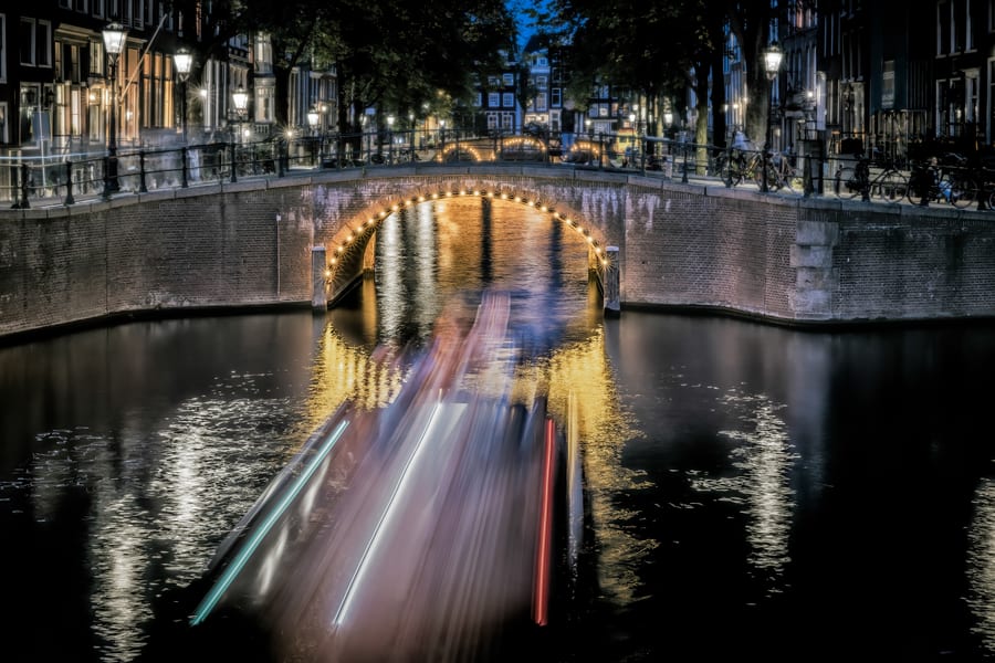 Avondfoto van een Amsterdamse gracht met brug en lichtsporen van een rondvaartboot